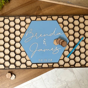 May include: A wooden hexagonal sign with a honeycomb pattern and a light blue hexagon in the center. The text "Brenda & James" is written in white cursive script on the blue hexagon. The date "04.19.24" is written below the names.