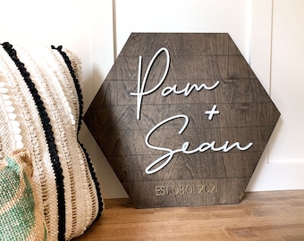 May include: A hexagonal wooden sign with the names "Pam + Sean" in white script and the date "EST 08 01 2021" in gold lettering.