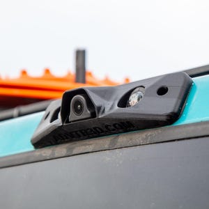 May include: A black rear-view camera mounted on the back of a teal car. The camera has a lens and a small light. The camera is labeled "ravtied.com".