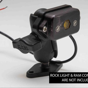 May include: Black RAM mount with a black and silver light fixture attached. The light fixture has two hexagonal buttons and a yellow lens. The text "ROCK LIGHT & RAM COMPONENTS ARE NOT INCLUDED" is printed below the mount.