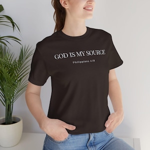 Puede incluir: Una camiseta marrón oscuro con el texto blanco "GOD IS MY SOURCE" y "Philippians 4:19". La camiseta es de cuello redondo y está hecha de un tejido suave.