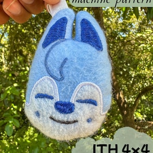 May include: A light blue felt ornament in the shape of a cartoon animal face with blue accents and white details. The text "Embroidery machine pattern" and "ITH 4x4 in the hoop" are visible. The ornament is held up against a green background.
