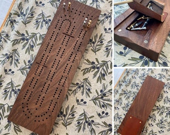 May include: A wooden cribbage board with a dark brown finish. The board has a track for pegs and a scoring area. The board is closed and has a leather flap on the side.