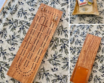 May include: A wooden cribbage board with a brown stain. The board has a track for pegs and a scoring area. The board is on a fabric background with a leaf pattern.
