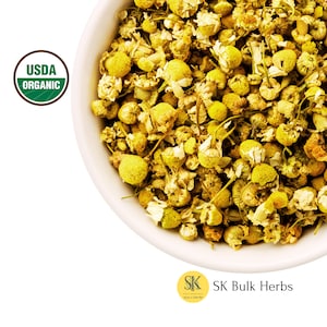May include: A white bowl filled with dried chamomile flowers. The flowers are a pale yellow colour and have a slightly brown tint. The bowl is sitting on a white background. The USDA Organic seal is visible in the top left corner of the image.