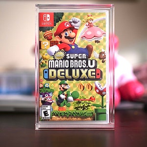 以下が含まれることがあります： 透明なプラスチックケースにビデオゲームカートリッジが入っています。ゲームはNintendo Switch用「New スーパーマリオブラザーズU デラックス」です。ケースの前面には、マリオ、ルイージ、ピーチ、キノピオが描かれています。ゲームは全年齢対象（E）です。
