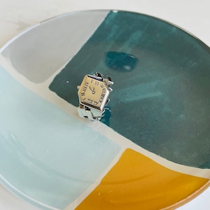 May include: A silver ring with a square face featuring Roman numeral clock hands. The ring is on a blue, green, and yellow plate.