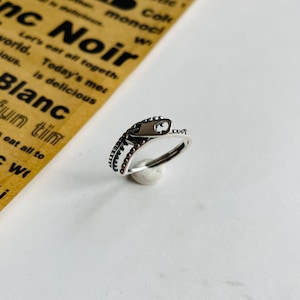 May include: A silver ring featuring a zipper design. The zipper has a small silver charm attached. The ring is displayed on a white surface.