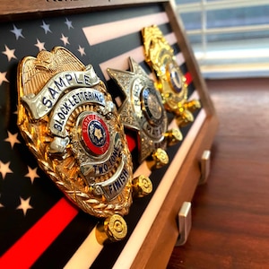 May include: A wooden shadow box display with an American flag background. Three gold and silver police badges are mounted on the display. The badges have the text "SAMPLE BLOCK LETTERING TWO TONE FINISH" on them.