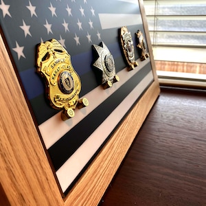 May include: A wooden frame with a thin blue line flag and four gold and silver badges. The badges are mounted on the flag with small screws. The frame is on a wooden surface.