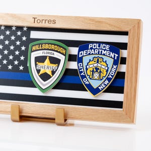 May include: A wooden frame containing a thin blue line flag graphic with the name Torres engraved on the top edge. Two police badges are mounted on the front: a green and black Hillsborough Florida Sheriff badge and a blue and gold NYPD badge.