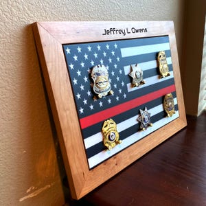 May include: A wooden framed display featuring an American flag design with a red line. The display holds several gold and silver badges. The name "Jeffrey L Owens" is printed at the top. The frame is made of light-colored wood.