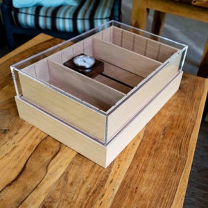 May include: A rectangular wooden humidor featuring a clear acrylic lid and multiple internal dividers. The interior contains a small, circular analog hygrometer mounted on a dark wooden block, designed for the organized storage of cigars.