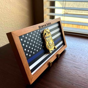 May include: A wooden shadow box with a thin brown frame displays a black and white American flag with a blue stripe. The flag is behind a gold badge with the text "Pinkerton Captain Security Systems". The name "Brianna Sebio" is engraved on the top of the frame.