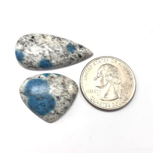 May include: Two polished k2 jasper stones with blue and white patterns, next to a US quarter coin. One stone is teardrop-shaped, the other is heart-shaped. The quarter coin is included for size reference.
