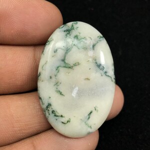 Pode incluir: Um caboch&atilde;o oval branco com inclus&otilde;es de &aacute;gata musgosa verde. A pedra &eacute; polida e tem uma superf&iacute;cie lisa.