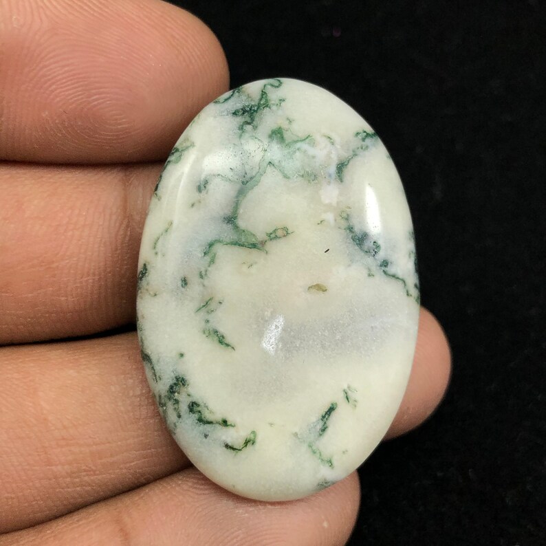 Pode incluir: Um caboch&atilde;o oval branco com inclus&otilde;es de &aacute;gata musgosa verde. A pedra &eacute; polida e tem uma superf&iacute;cie lisa.