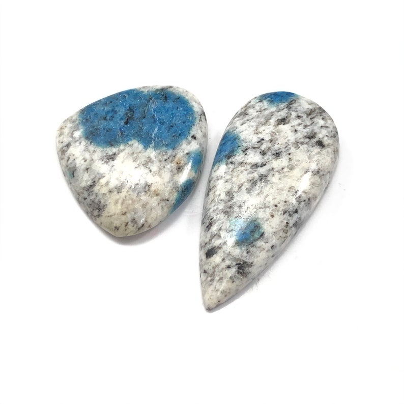 May include: Two polished k2 jasper stones. One is a rounded triangle shape, the other is a teardrop shape. Both stones have a white base with black speckles and patches of blue.