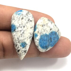 May include: Two polished k2 jasper gemstones. One is teardrop-shaped, the other is a rounded triangle. Both stones have a white base with black speckles and patches of blue.