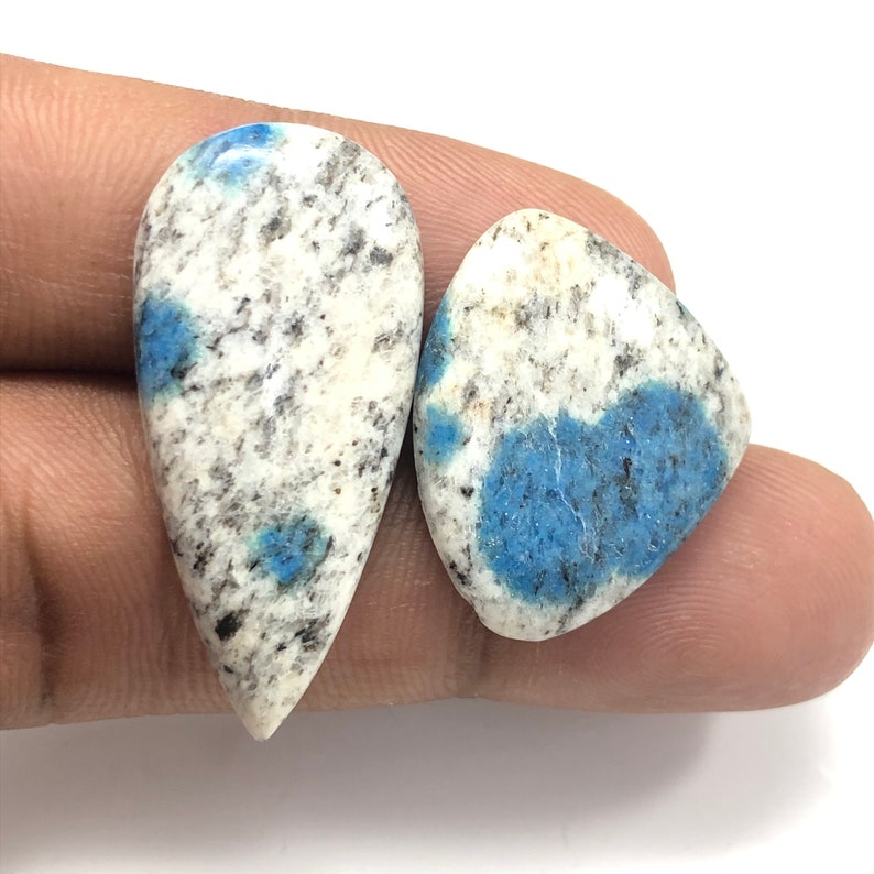May include: Two polished k2 jasper gemstones. One is teardrop-shaped, the other is a rounded triangle. Both stones have a white base with black speckles and patches of blue.
