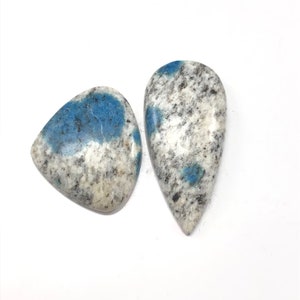 May include: Two K2 Azurite granite gemstones. One is a rounded triangle shape, the other is a teardrop shape. Both stones have a white base with black speckles and patches of blue.