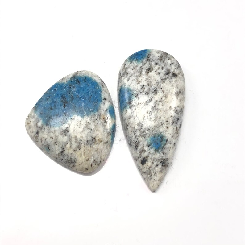 May include: Two K2 Azurite granite gemstones. One is a rounded triangle shape, the other is a teardrop shape. Both stones have a white base with black speckles and patches of blue.