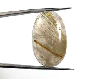 May include: A clear oval-shaped gemstone with gold inclusions. The gemstone is held in tweezers against a white background.