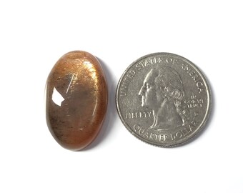 May include: A large, oval-shaped sunstone cabochon with a golden-brown color and a shimmery, iridescent effect. The stone is approximately the size of a US quarter.