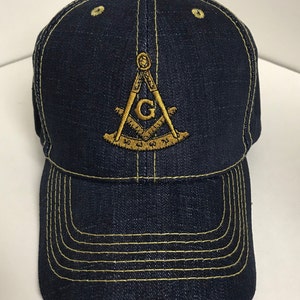 May include: A dark blue denim baseball cap with a gold embroidered Masonic symbol on the front. The symbol includes a compass, square, and the letter 'G'.