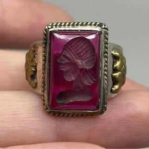 Puede incluir: Un anillo vintage con una piedra rectangular de color rojo intenso que presenta un perfil grabado de una persona. El anillo tiene una banda plateada con detalles dorados en los lados y un borde decorativo alrededor de la piedra. El anillo está sostenido por los dedos de una persona.