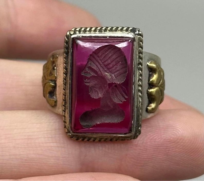 K&ouml;nnte beinhalten: Ein Vintage-Ring mit einem rechteckigen, tiefroten Stein, der ein eingraviertes Profil einer Person zeigt. Der Ring hat ein silbernes Band mit goldfarbenen Akzenten an den Seiten und eine dekorative Bord&uuml;re um den Stein. Der Ring wird von den Fingern einer Person gehalten.