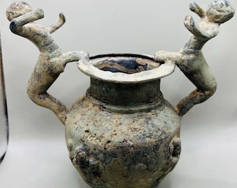 Pode incluir: Um vaso antigo de bronze desgastado com duas alças, cada uma adornada com uma pequena figura humana estilizada. O vaso tem um corpo arredondado e uma borda larga e ligeiramente alargada. O bronze tem uma aparência manchada e envelhecida.