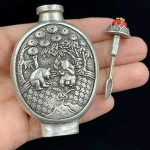 May include: A silver-colored, oval-shaped snuff bottle with a detailed relief scene of pandas and bamboo. The bottle has a matching stopper with a red gemstone and a small spoon. The design is intricate and the bottle is held in a hand.