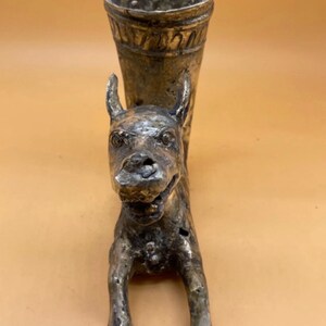 Puede incluir: Un jarrón de metal plateado con forma de perro. El jarrón tiene una parte superior acampanada con una banda decorativa y una cabeza y cuerpo de perro detallados. El perro está de pie, con las patas delanteras extendidas.