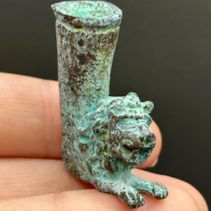May include: A small, antique bronze sculpture of a lion. The lion is lying down with its front paws extended. The sculpture has a cylindrical tube extending upwards from the lion's back. The bronze has a green patina.
