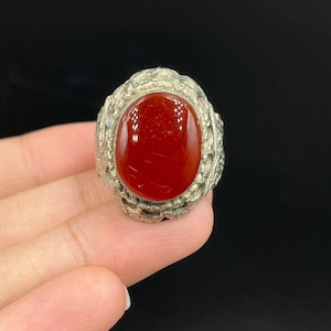 Puede incluir: Anillo de plata con una piedra ovalada de color rojo intenso. El engaste del anillo presenta un diseño texturizado y ornamentado. La piedra pulida refleja la luz, sobre un fondo oscuro.