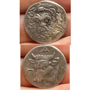 May include: Two silver antique coins. The top coin features a detailed profile of a bearded figure. The bottom coin displays a bull's head with the letter "A" above it. Both coins show signs of wear and age.