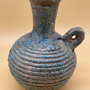 May include: A small, blue and gray ceramic jug with a round body and a narrow neck. The jug has a handle with a hole and is decorated with horizontal ridges. The color is a mottled blue and gray, giving it a rustic appearance. The jug is likely handmade.