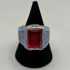 May include: A silver ring with a large, rectangular, red gemstone. The ring band is blue with red, green, and white decorative accents. The ring is displayed on a black velvet stand.