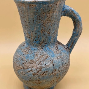 May include: A handcrafted, textured ceramic pitcher with a bulbous base and a flared top. The pitcher features a blue and brown mottled glaze, with a curved handle. The overall design is rustic and artistic, suitable for decorative use.
