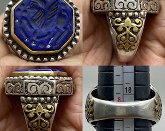 Può includere: Un anello d'argento con una grande pietra blu scuro ottagonale. La pietra è incastonata in una lunetta dorata e presenta un disegno scolpito. L'anello ha dettagli argentati e dorati ornati sulla fascia.
