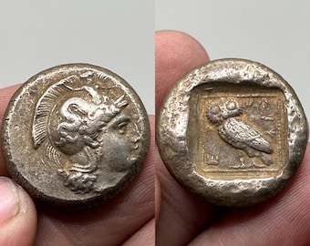 May include: Two silver ancient Greek coins. One coin features a profile of a helmeted Athena. The other coin shows an owl within a square frame. Both coins have a worn, aged appearance.