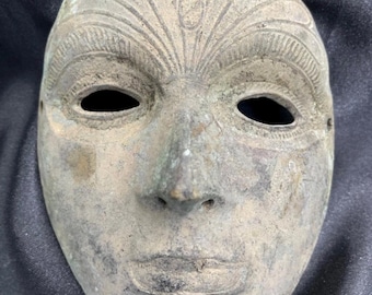 May include: A weathered, antique-style mask with a serene expression. The mask features open eye sockets, a defined nose, and a slightly parted mouth. The top of the mask has a decorative, fan-like design. The mask is a muted, earthy tone.