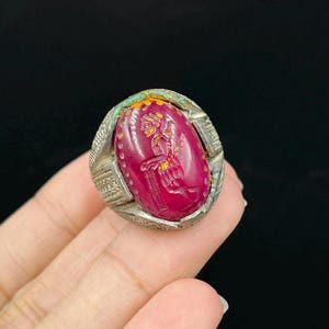 Op de afbeelding: Een zilveren ring met een ovale, dieprode steen. De steen heeft een gebeeldhouwde figuur met gouden en gele accenten. De ring heeft ingewikkelde zilveren details rond de steen en de band. De ring wordt door een persoon vastgehouden.