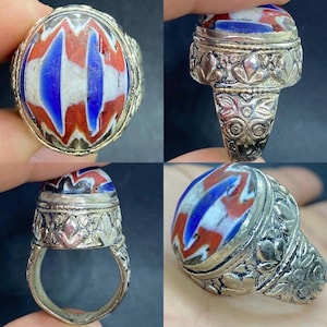 May include: A silver ring with an oval-shaped, colorful glass stone. The stone features a pattern of red, white, and blue stripes. The ring's band and setting are detailed with floral designs, showcasing an antique aesthetic.
