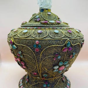 May include: Ornate antique brass container with a filigree design, adorned with colorful gemstones. The container has a domed lid topped with a clear, crown-shaped finial. The base is round and decorated with more gemstones.