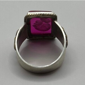 K&ouml;nnte beinhalten: Silberring mit einem rechteckigen, magentafarbenen Stein. Der Stein ist mit einem floralen Design graviert. Der Ring hat ein dekoratives Band mit goldfarbenen Akzenten. Ein Schmuckst&uuml;ck.