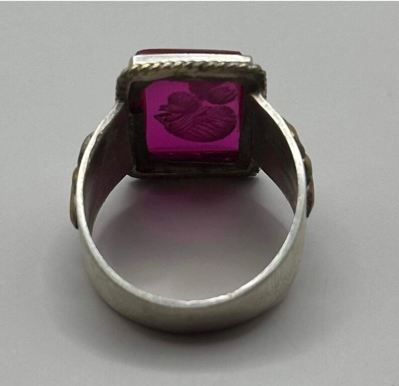 K&ouml;nnte beinhalten: Silberring mit einem rechteckigen, magentafarbenen Stein. Der Stein ist mit einem floralen Design graviert. Der Ring hat ein dekoratives Band mit goldfarbenen Akzenten. Ein Schmuckst&uuml;ck.