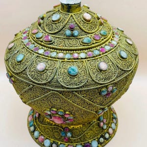 Puede incluir: Contenedor ornamentado de estilo antiguo con un diseño de filigrana en tonos dorados. El contenedor está adornado con piedras preciosas de colores en tonos rosa, azul y verde. Una piedra transparente y redondeada corona la tapa.