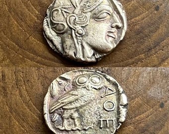 May include: A silver coin featuring a profile of a woman's face with detailed hair and headwear. The reverse side shows an owl with intricate details, along with Greek lettering and symbols. The coin has a worn, antique appearance.